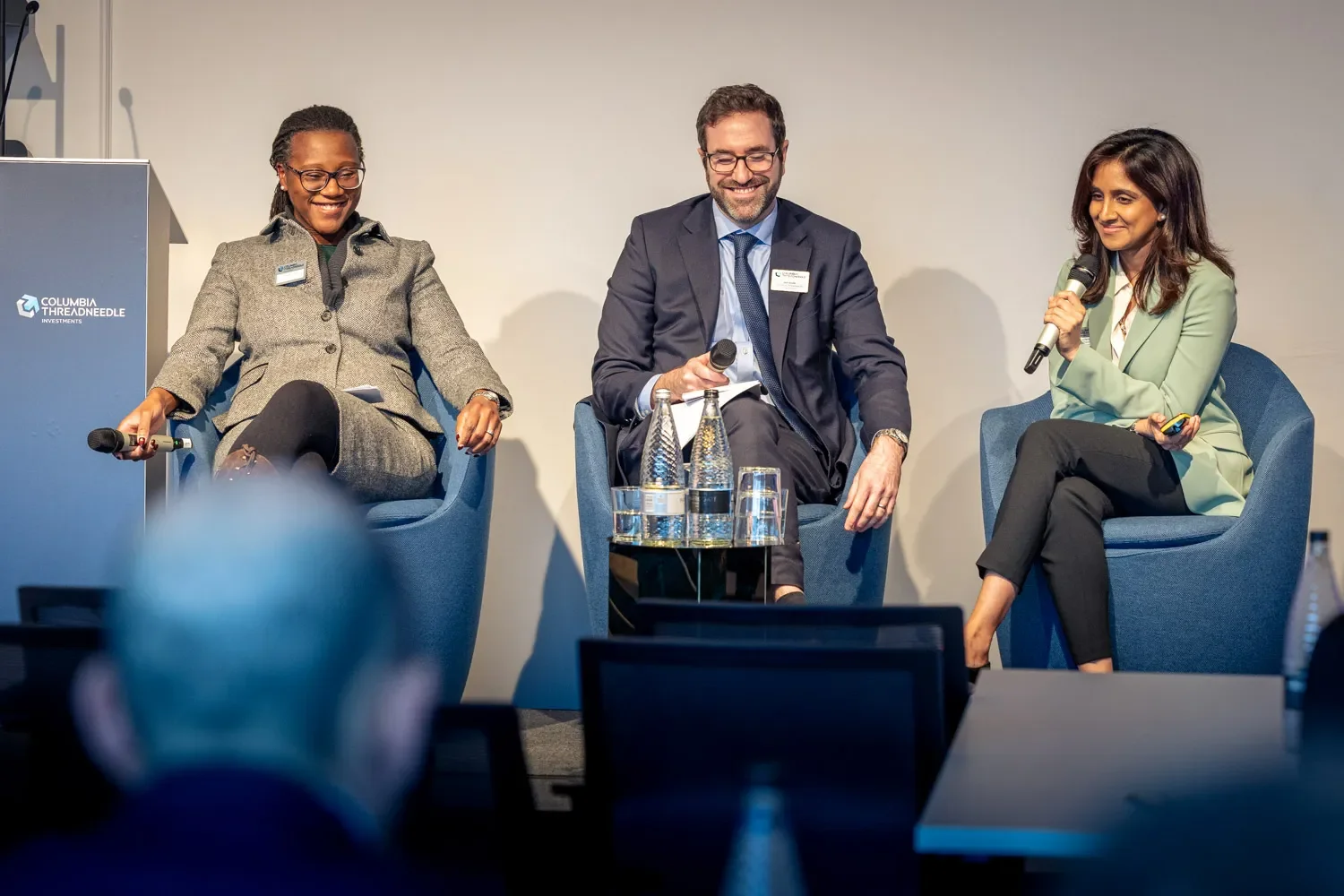 Three conference delegates are sitting on chairs, engaged in a conversation. They are wearing suits and ties, and one of them is holding a microphone. London event.