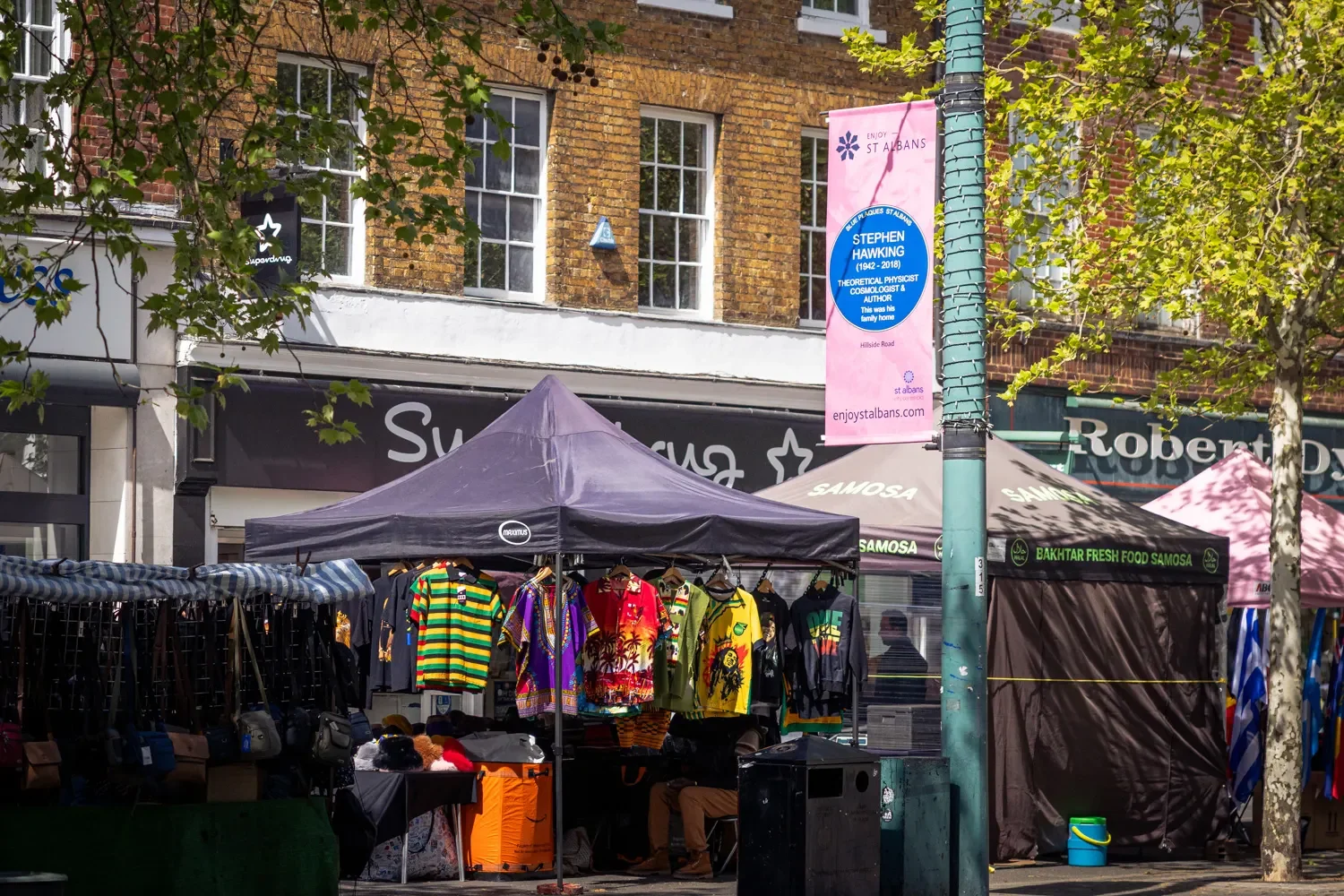St Albans BID banner featuring a Stephen Hawking blue plaque sign, mounted on a lamppost above busy market stalls in St Albans town centre