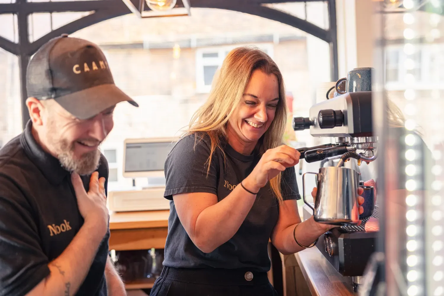 Two Nook café owners laughing together behind the espresso machine during their brand photography session in Markyate