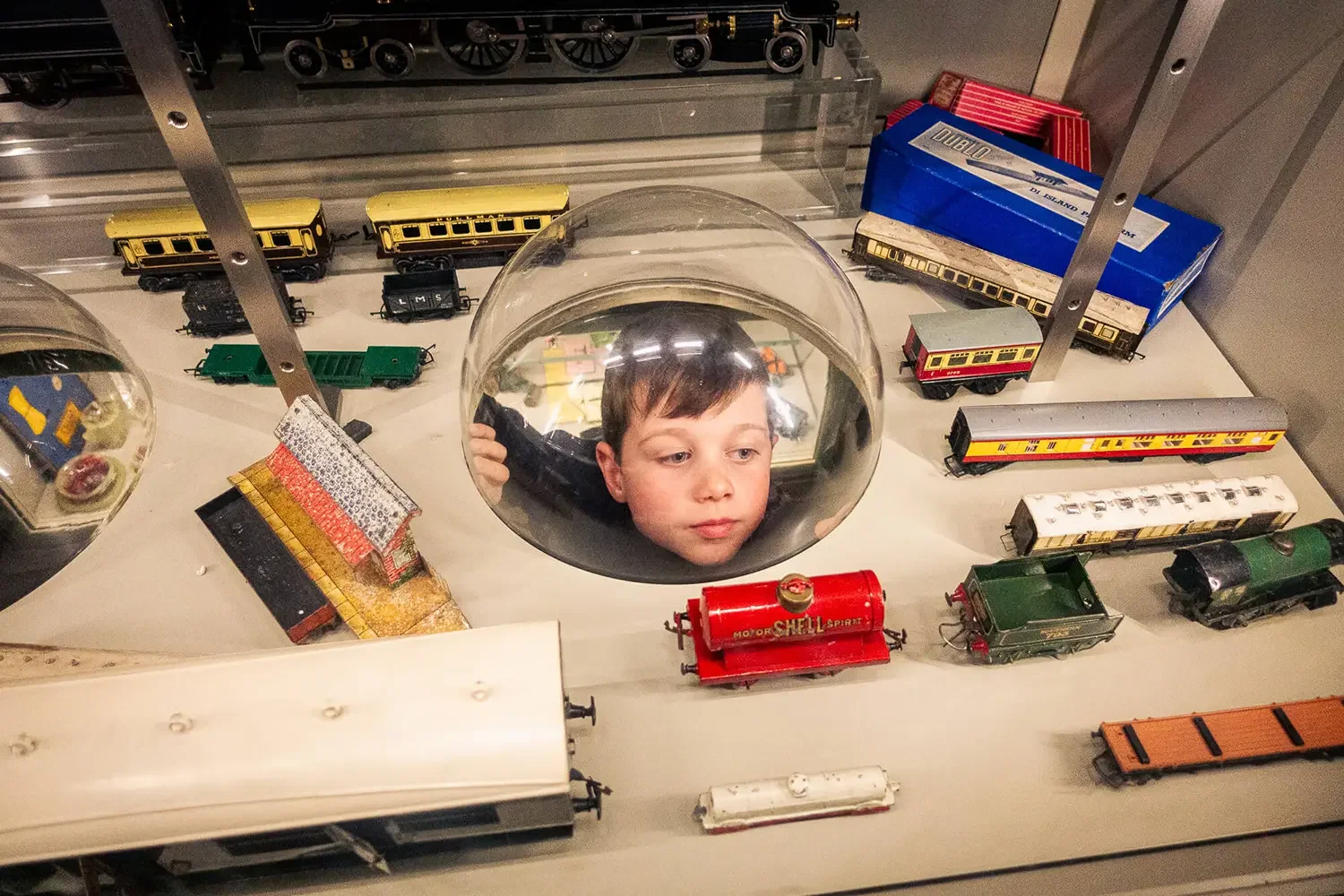 A small boy looking through an observation bubble at a miniature railway set.