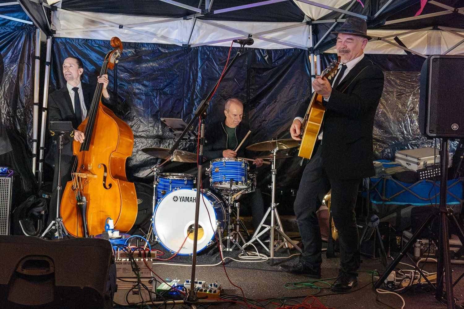 Five-piece jazz band in suits playing keyboard, saxophone, double bass, drums and guitar under a bunting-covered gazebo