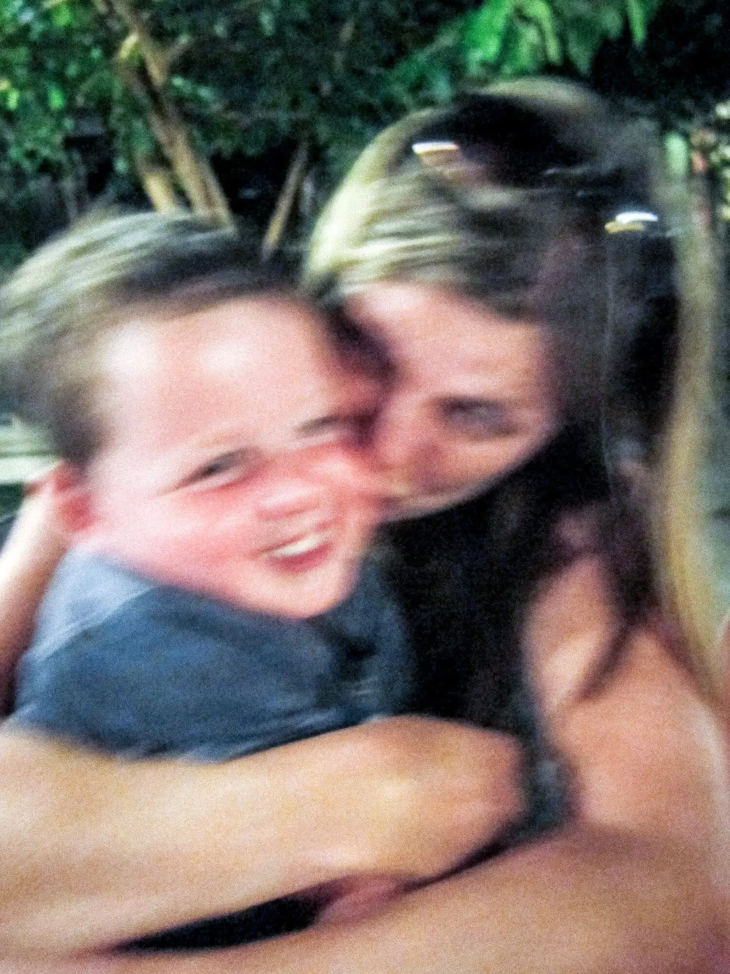 Two people hugging each other closely, smiling and appearing joyful, with a blurred background of greenery.