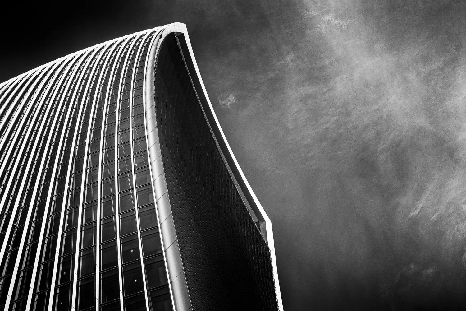 The sweeping curved glass facade of 20 Fenchurch Street rising against a dramatic cloudy sky, shot from directly below, black and white