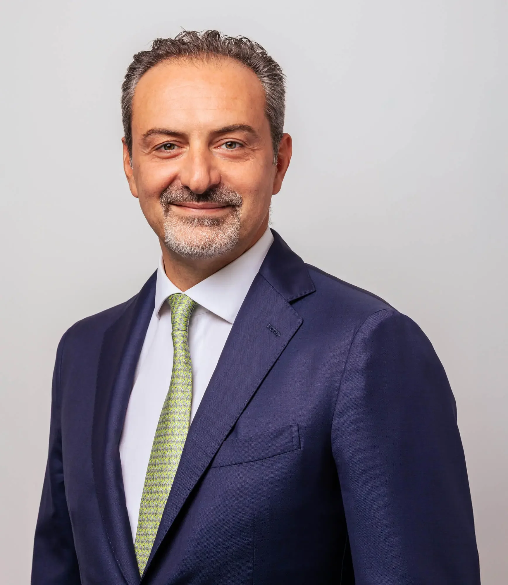 Headshot of a middle-aged man in a navy suit, white shirt, and pale green tie, smiling against a plain gray background.