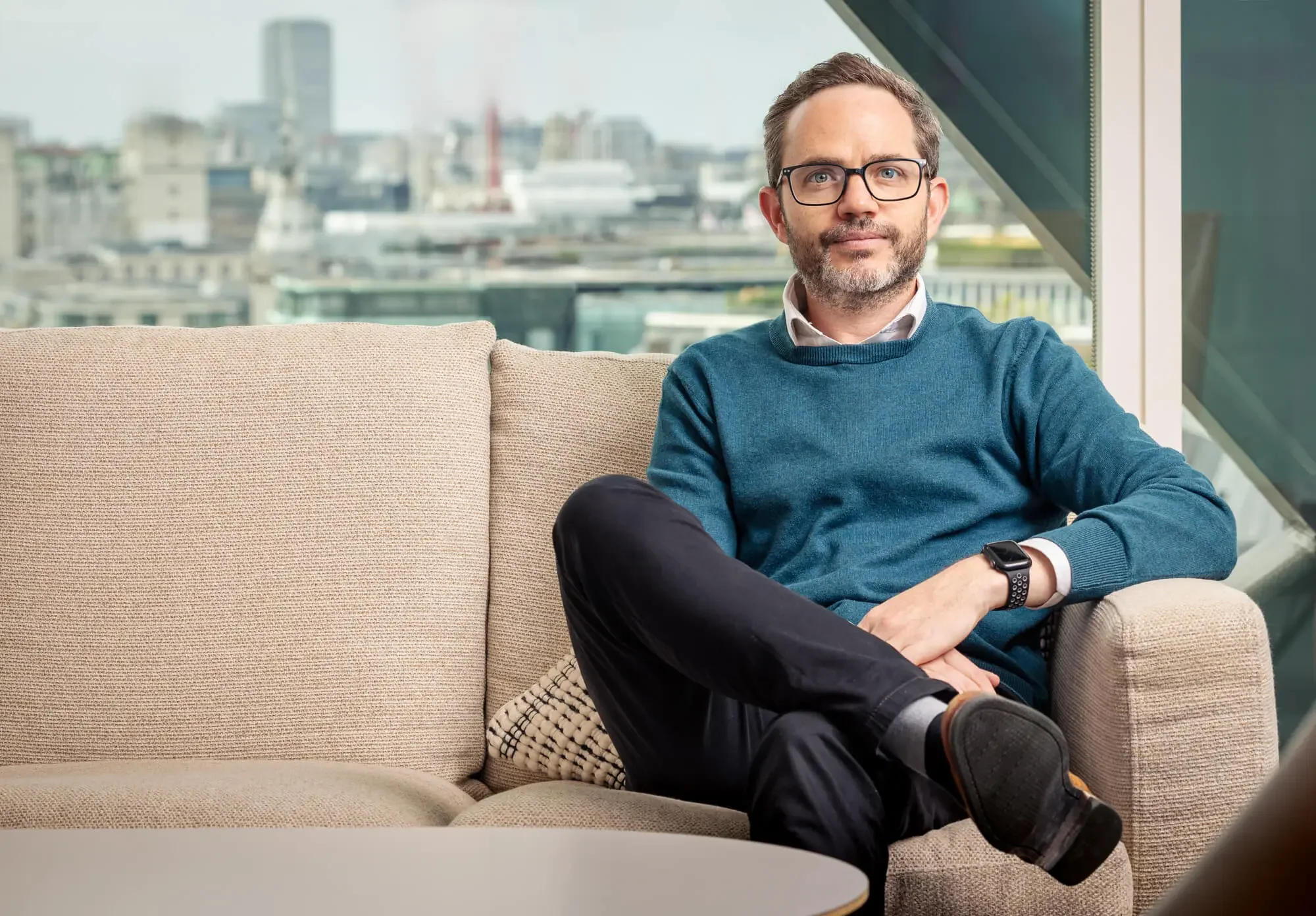 A man with glasses and a beard sitting on a beige sofa in a modern office lounge, with large windows showing city buildings in the background. He is wearing a blue sweater, white shirt, black pants, and a smartwatch.