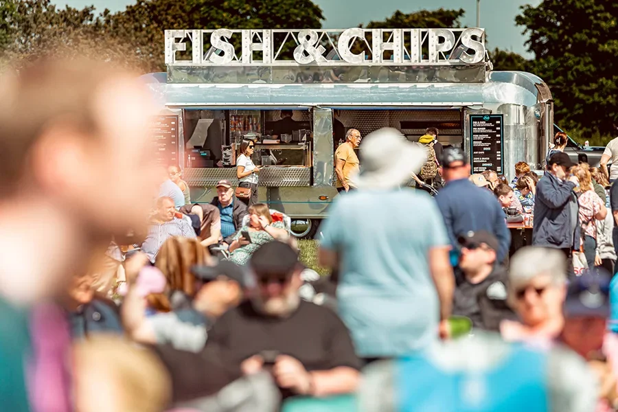 Crowded outdoor gathering with a fish and chips food truck under a sunny sky.
