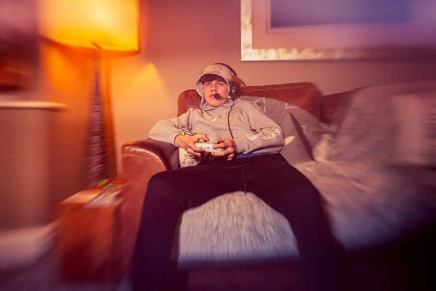 A teenage boy in a grey hoodie and gaming headset sits back in a leather armchair, white controller in hand, eyes fixed on the screen, bathed in warm amber lamplight.