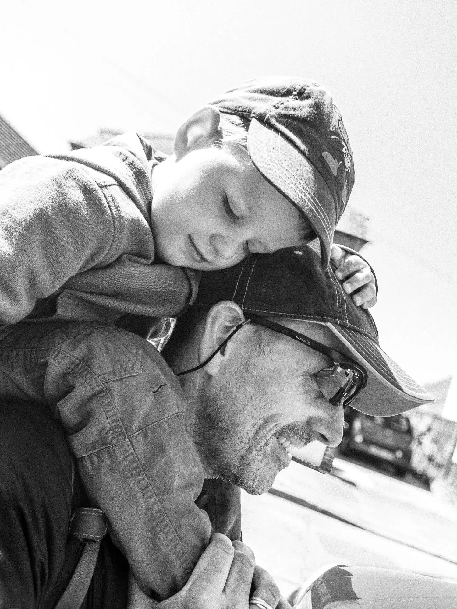 A young boy resting his head on an adult's shoulder, both wearing hats and sunglasses, outdoors on a sunny day.