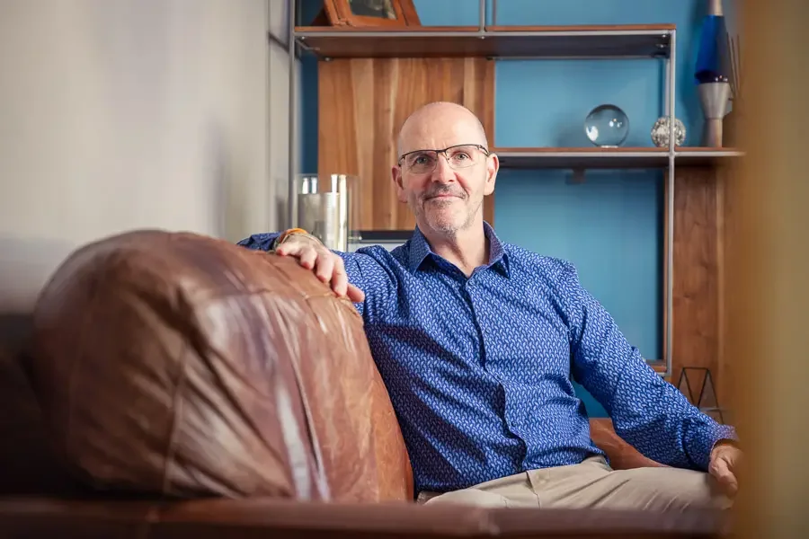 Self-portrait of Mike Dick from Mike Dick Photography, wearing a blue patterned shirt and beige trousers, sitting on a brown leather couch, resting his hand on the armrest, in a modern living room with wooden and blue decor.