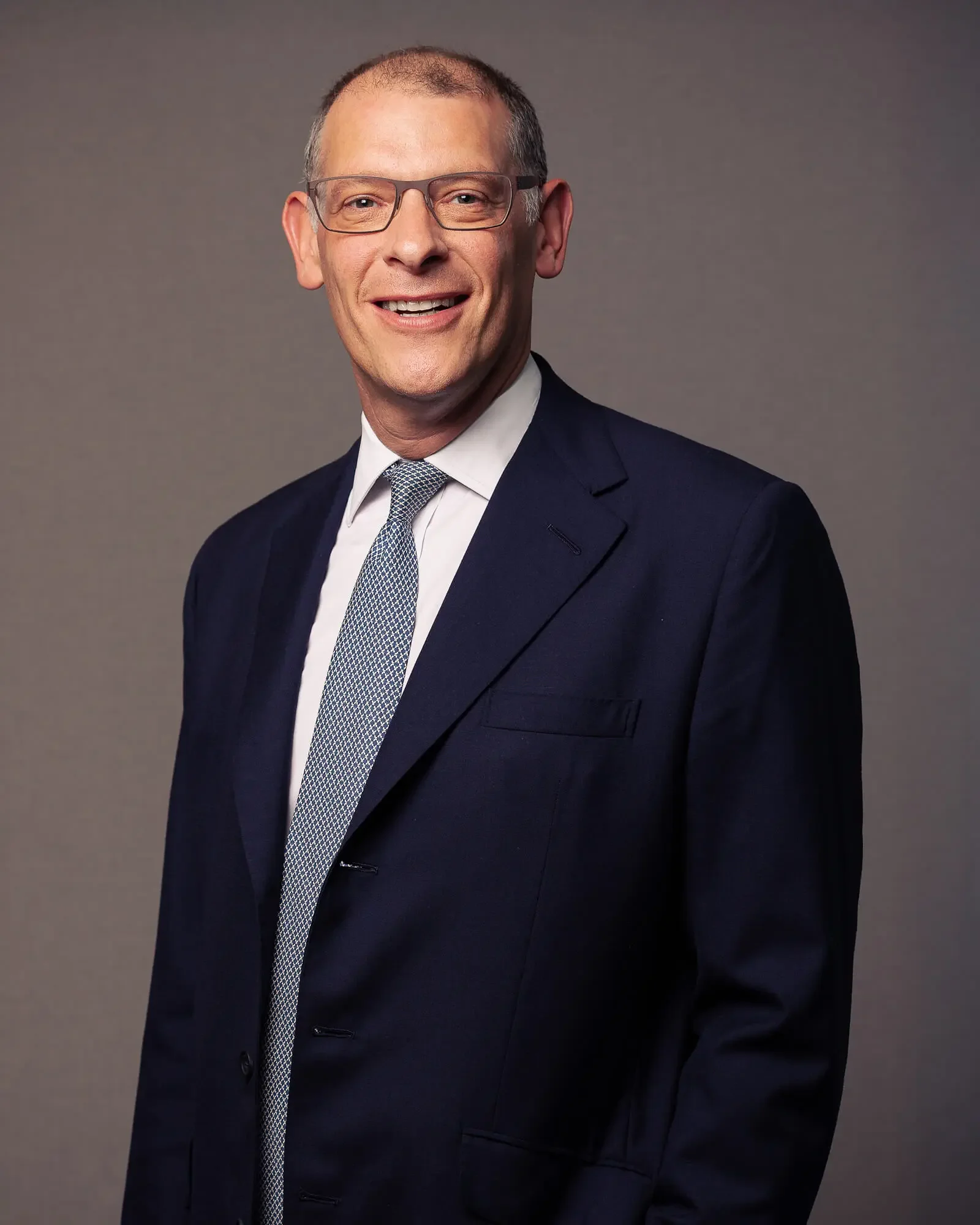 Portrait of a smiling man wearing glasses, a navy blue suit, white shirt, and a patterned tie, standing against a gray background.