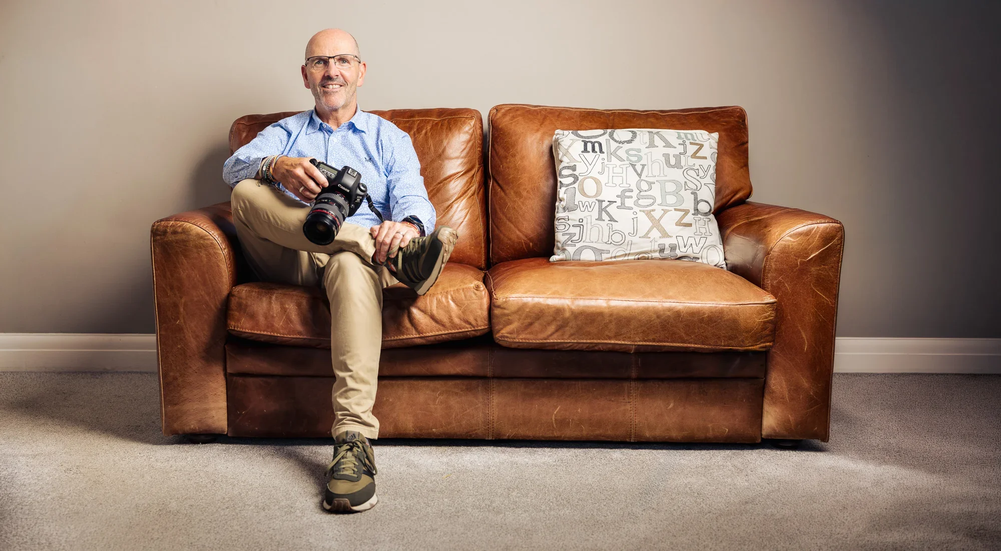 A middle-aged man with glasses and a beard sitting on a brown leather sofa, holding a professional camera, smiling, against a plain beige wall.
