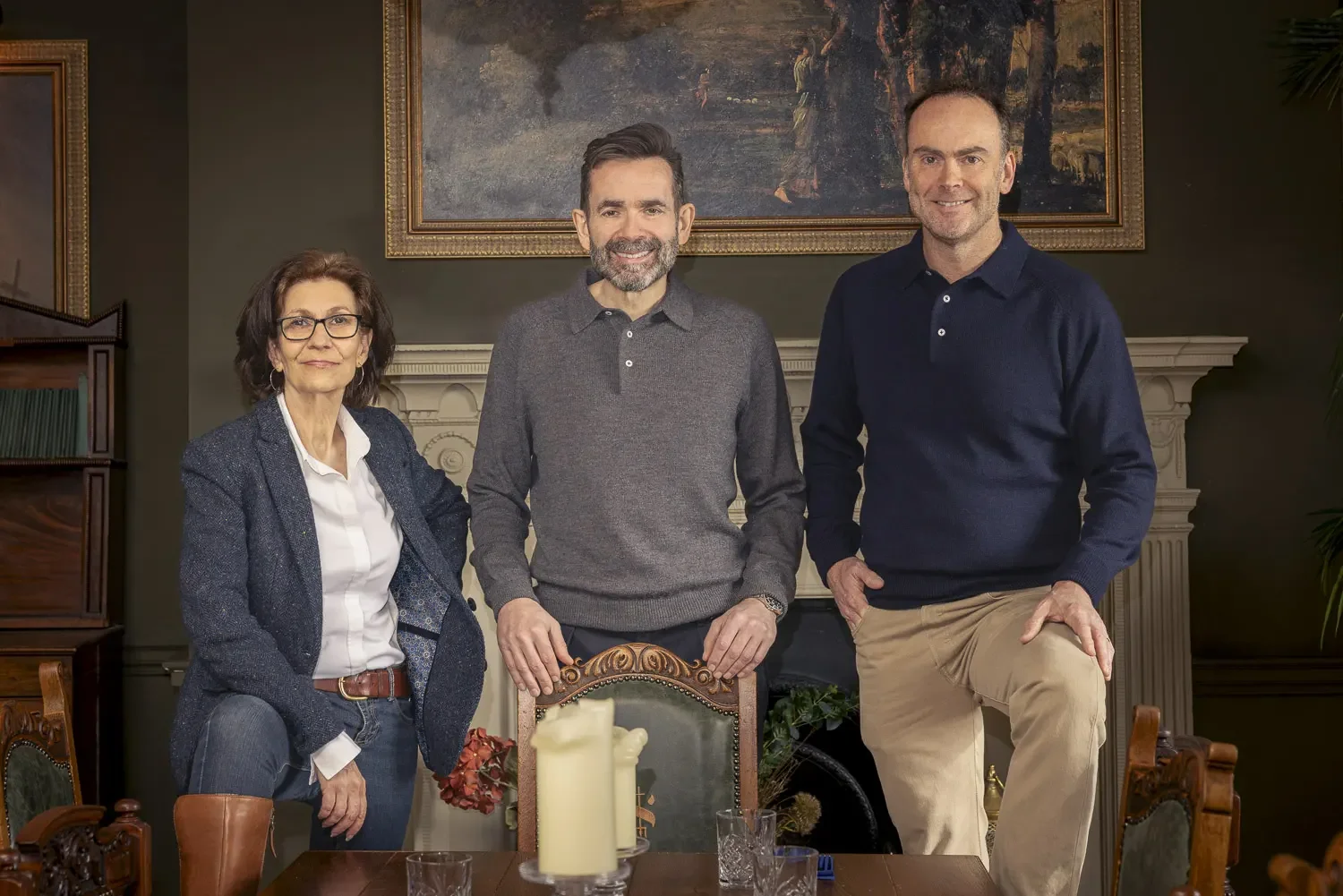 Three people standing in a room with dark green walls, in front of a fireplace with a painting above it. A woman on the left wearing glasses, a white shirt, a dark blazer, and boots. A man in the middle with dark hair and beard, wearing a gray sweate