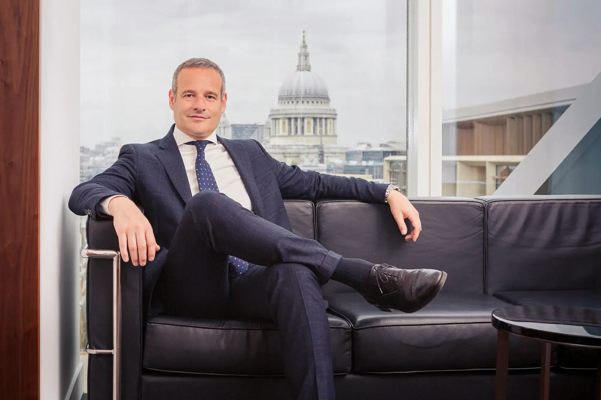 A man wearing a dark blue suit, white shirt, and navy polka dot tie sitting on a black leather couch with his legs crossed, inside a modern office with large windows and a cityscape view, including the dome of St. Paul's Cathedral.