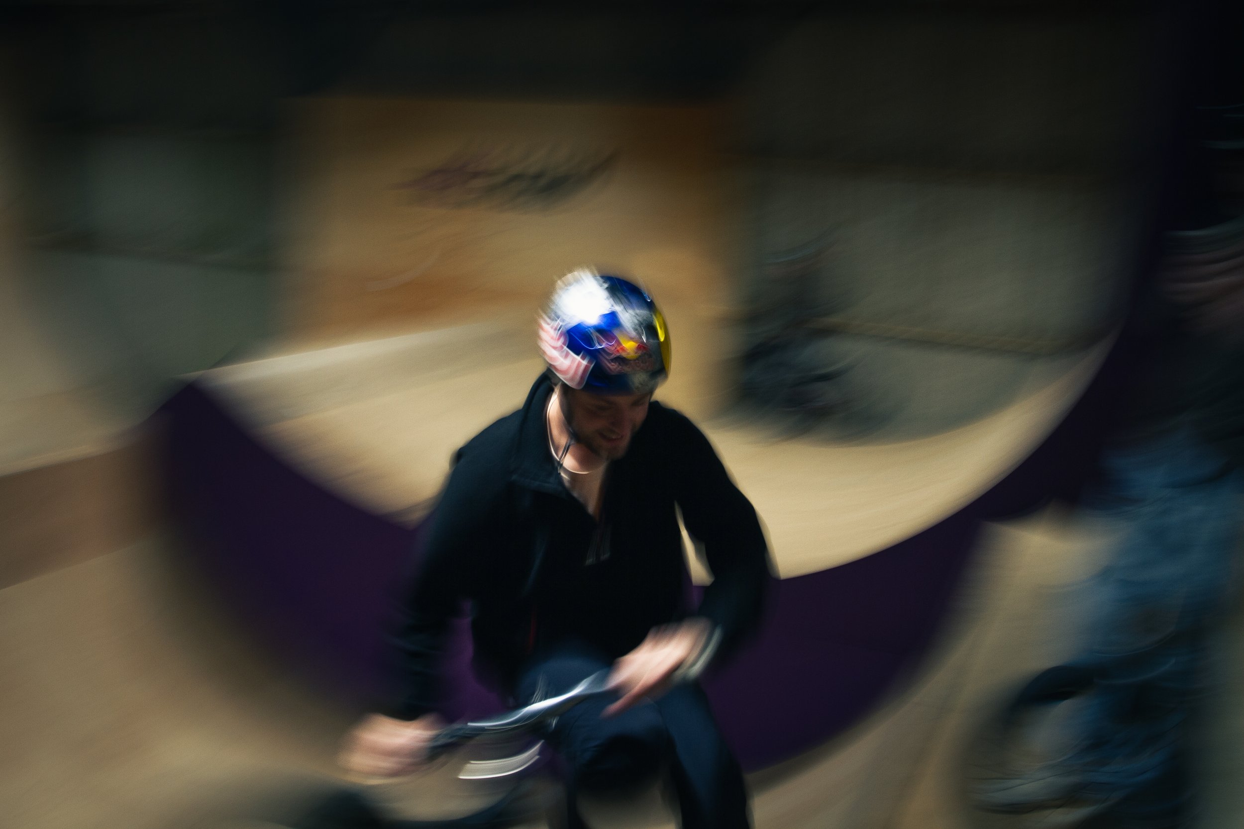 A person wearing a helmet is riding a bicycle in an indoor skatepark with a blurred background.