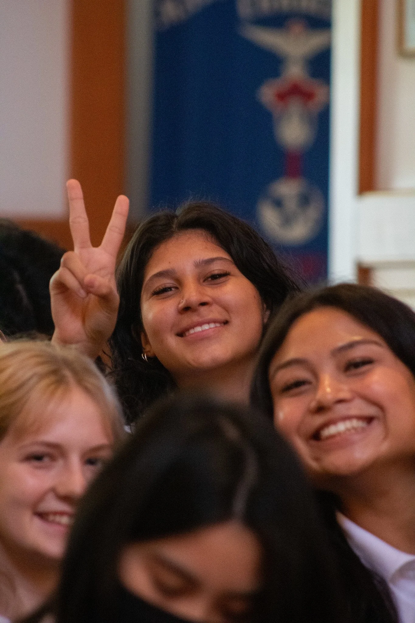 Several women smiling, with one woman making a peace sign with her fingers behind another woman's head.