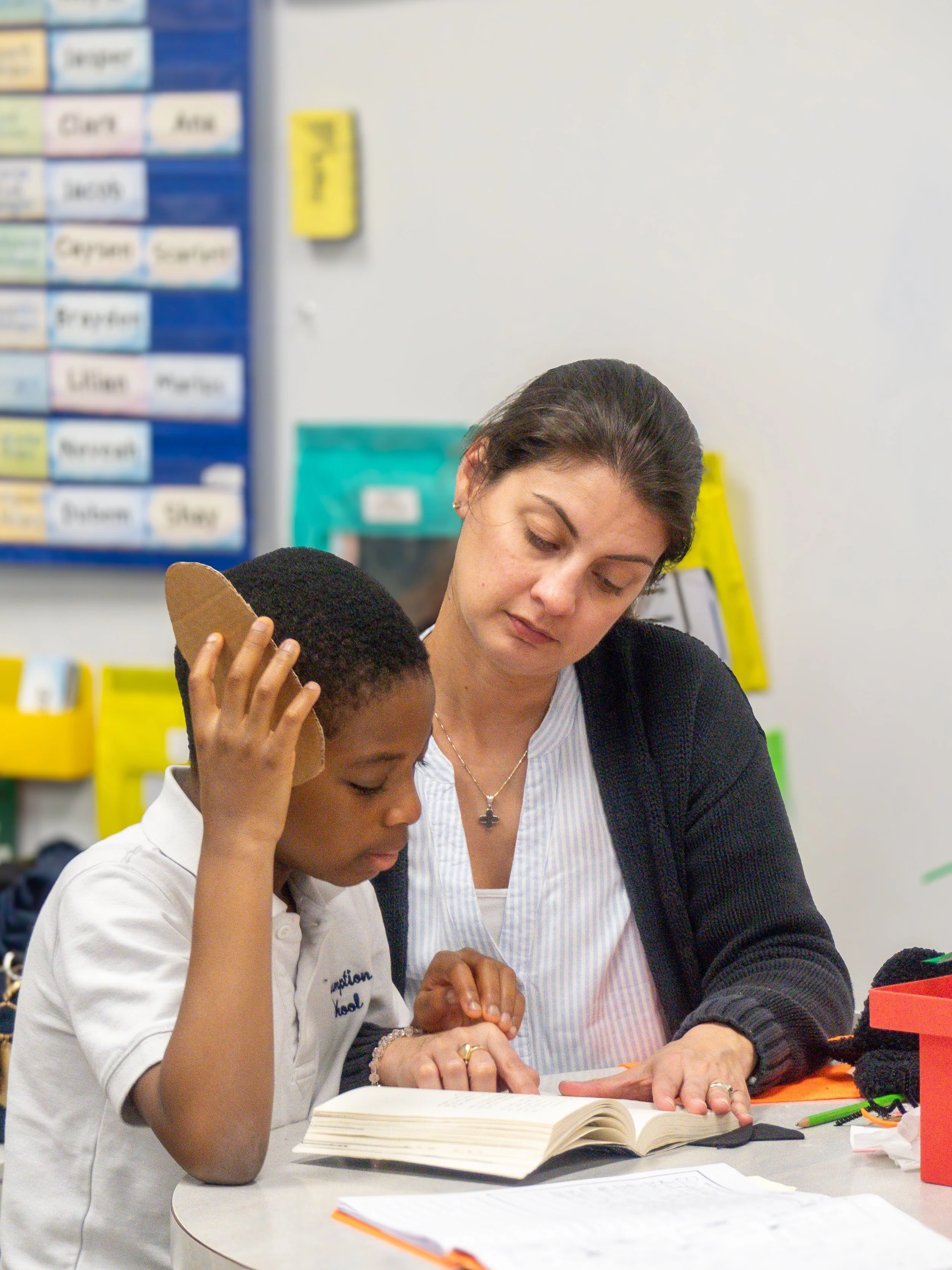 A woman and a boy are sitting at a table, reading a book together in a classroom. The boy is holding his head and looking at the book, while the woman is pointing at the pages.