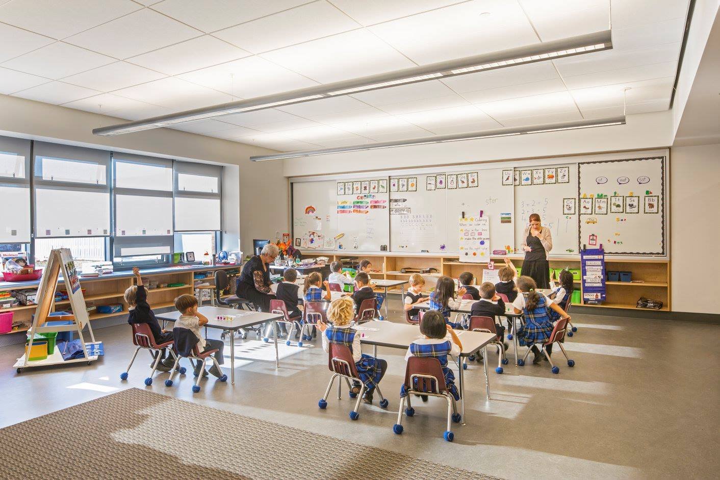 First grade classroom with students sitting at desks, teacher at whiteboard, and teacher's aide at side.