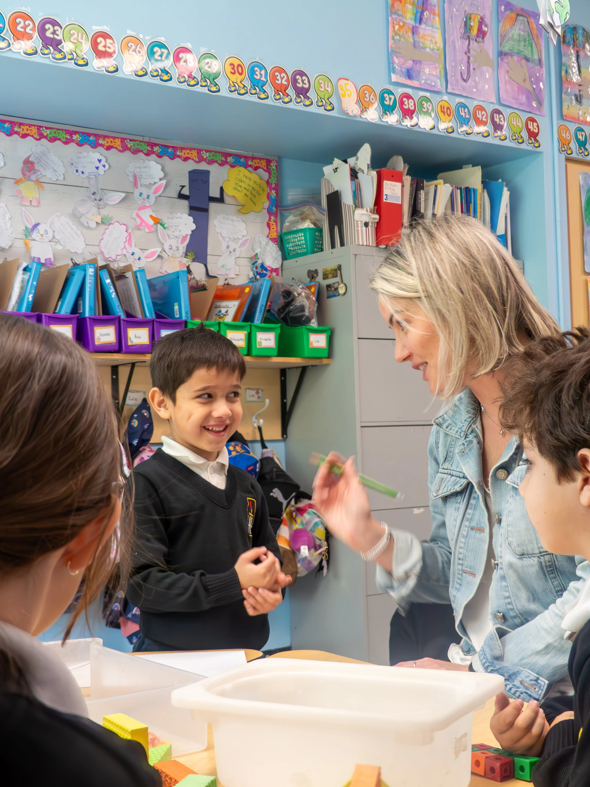 A classroom scene with a teacher smiling and talking to a group of students gathered around a table, with educational decorations and supplies in the background.