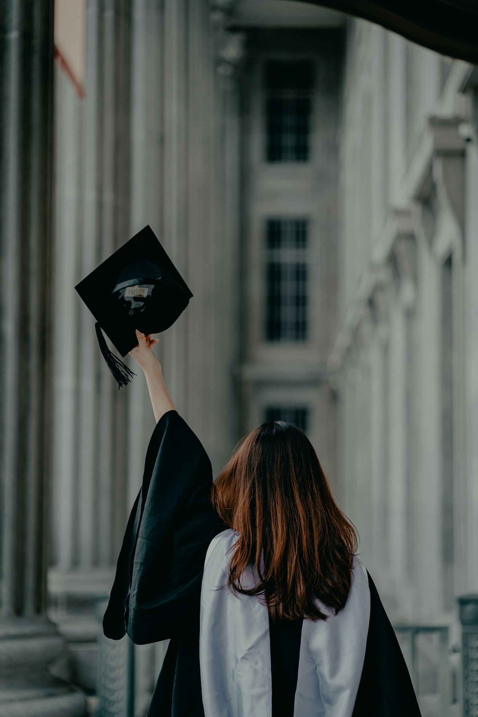 Graduate in cap and gown raising mortarboard in front of columned building.