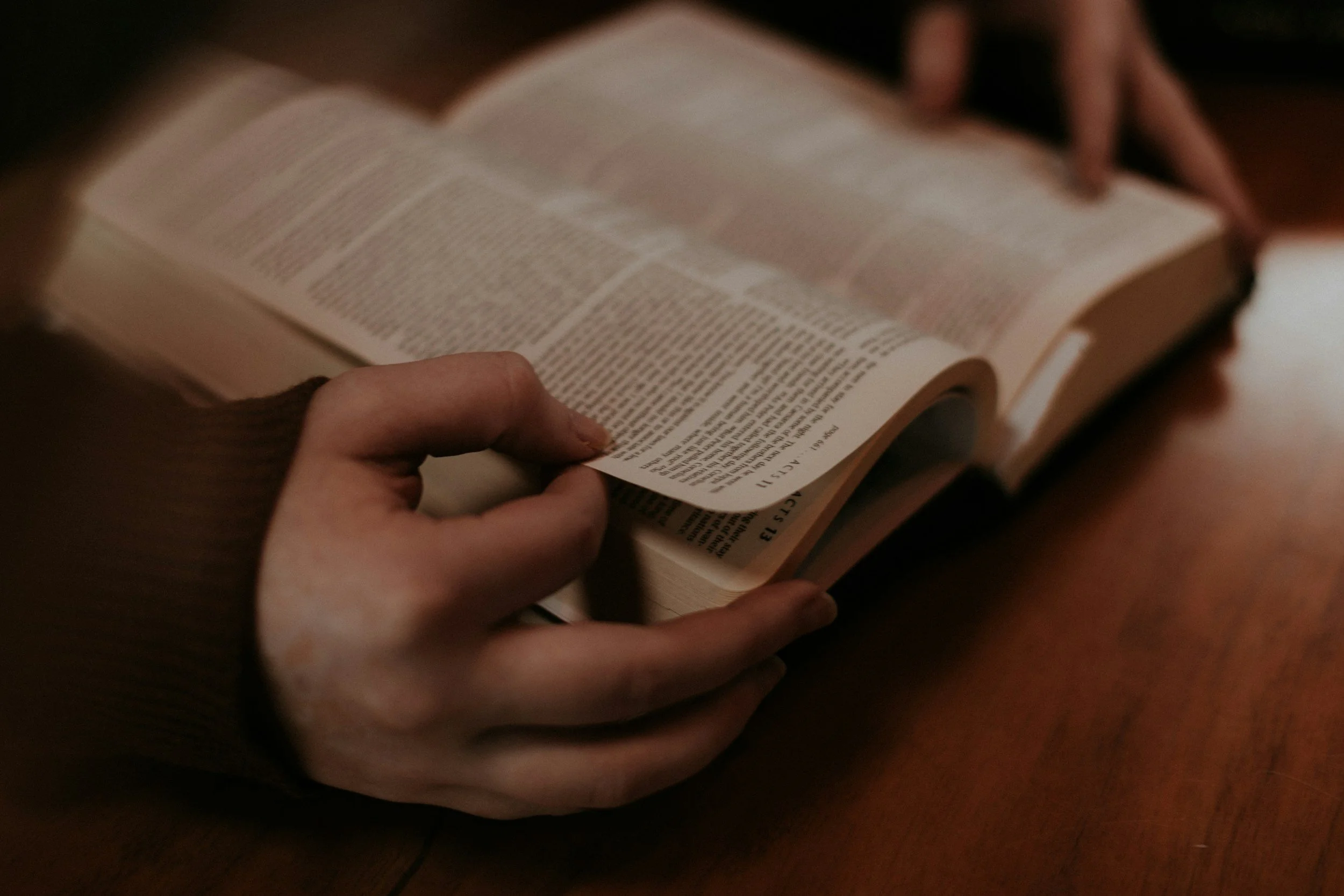 Person flipping through pages of an open Bible on a wooden table.