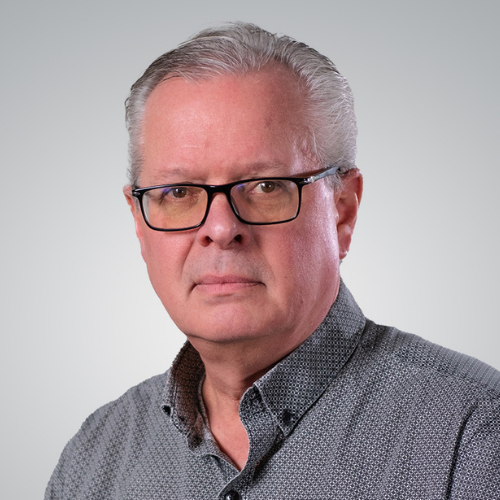 A middle-aged man with gray hair and glasses wearing a gray, patterned collared shirt poses for a portrait against a neutral background.