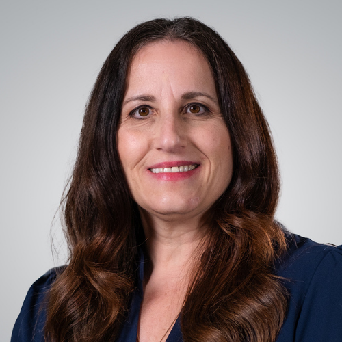 A woman with long brown hair wearing a navy blue top, smiling at the camera against a plain light gray background.