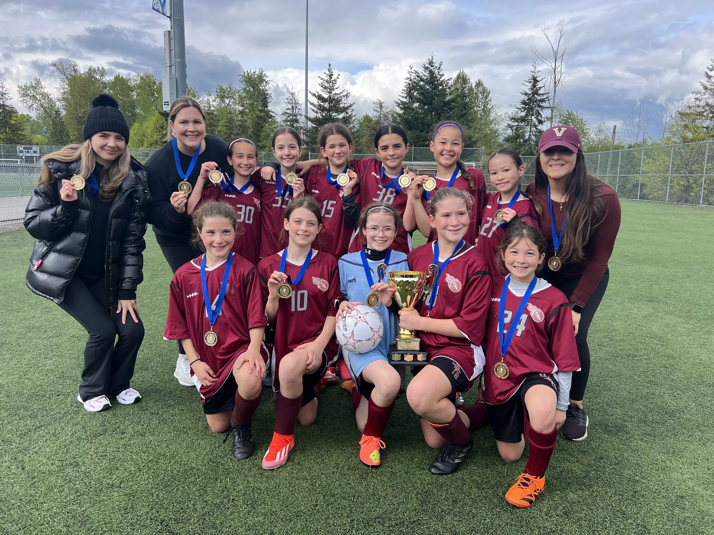 Youth girls soccer team in maroon uniforms celebrating with medals and a trophy on the field.