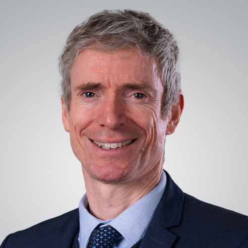 Portrait of a middle-aged man with gray hair, wearing a navy suit, light blue shirt, and blue tie, smiling against a plain light background.