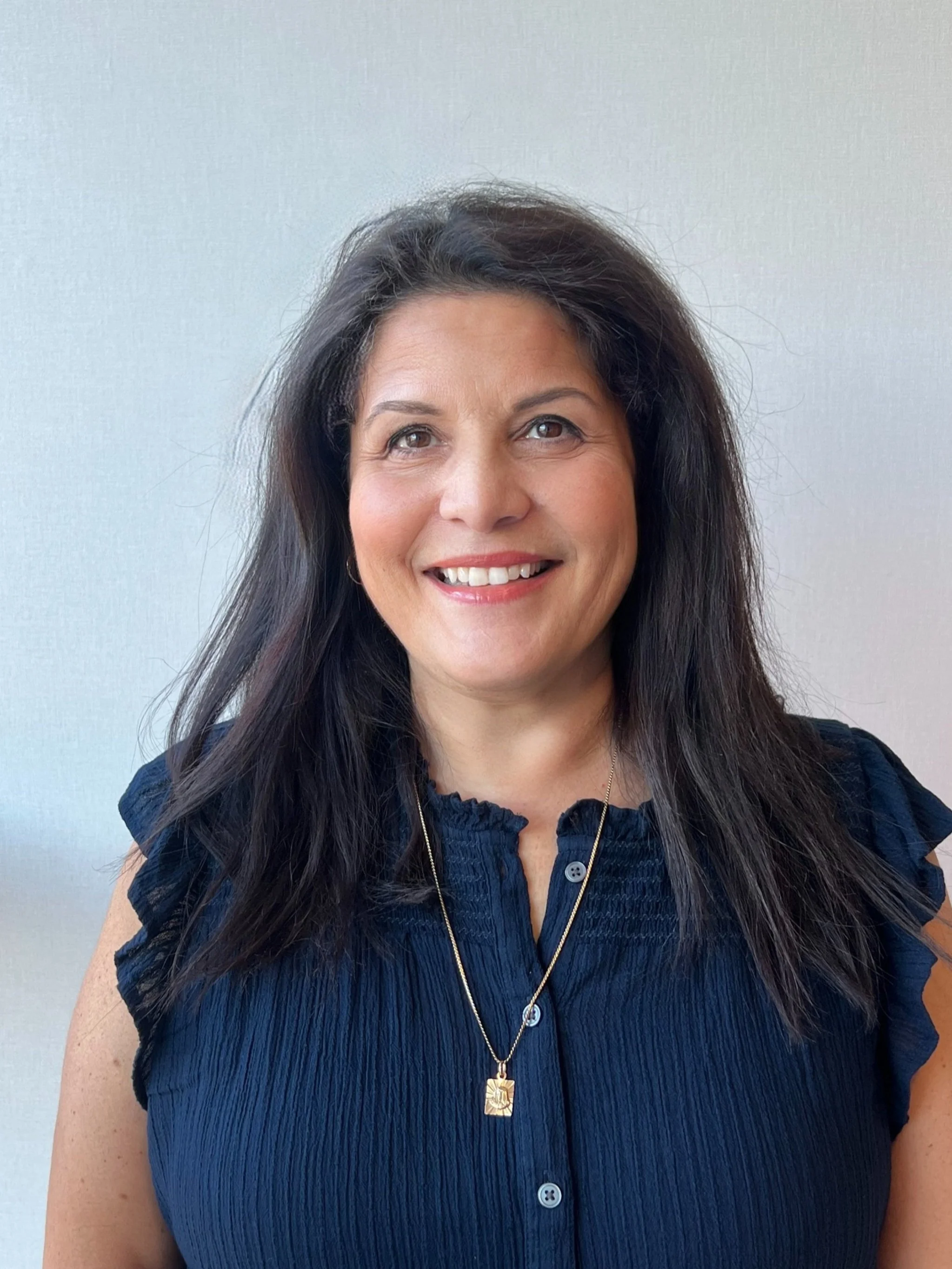 Portrait of a woman with long dark hair wearing a navy blue blouse and gold necklace, smiling against a plain light-colored background.