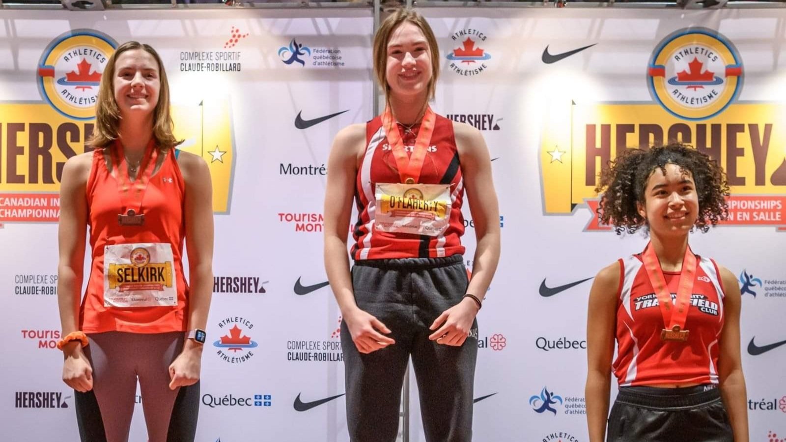 Three female athletes standing on a winners' podium in front of a backdrop with logos, wearing medals and red athletic uniforms. The woman in the center is in first place, flanked by the second and third place winners.