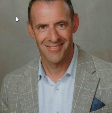 A man with short dark hair, wearing a light blue shirt and a checked gray blazer, smiling at the camera.