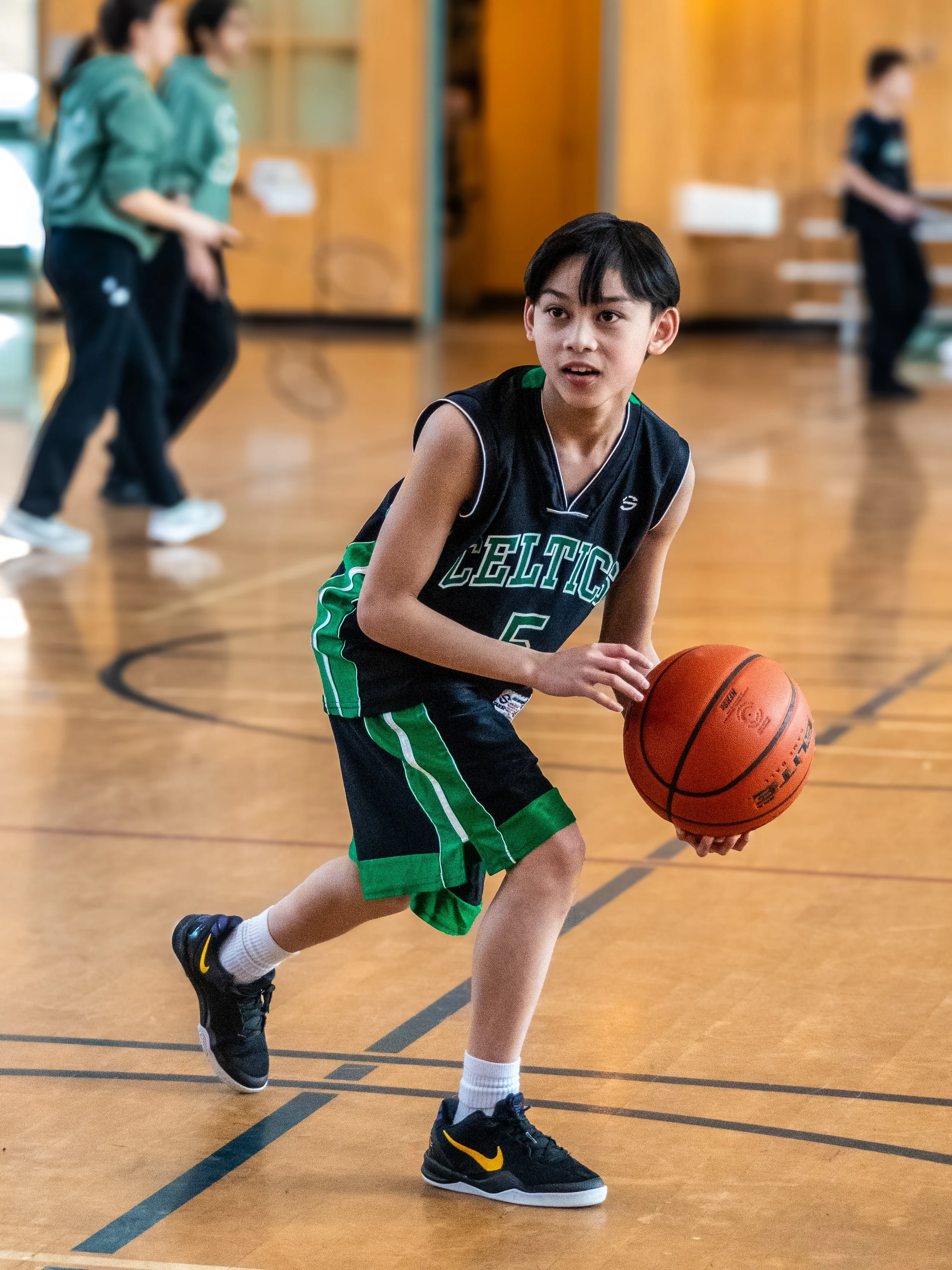 Young boy in a black and green basketball uniform dribbling a basketball on an indoor court with other kids playing in the background.