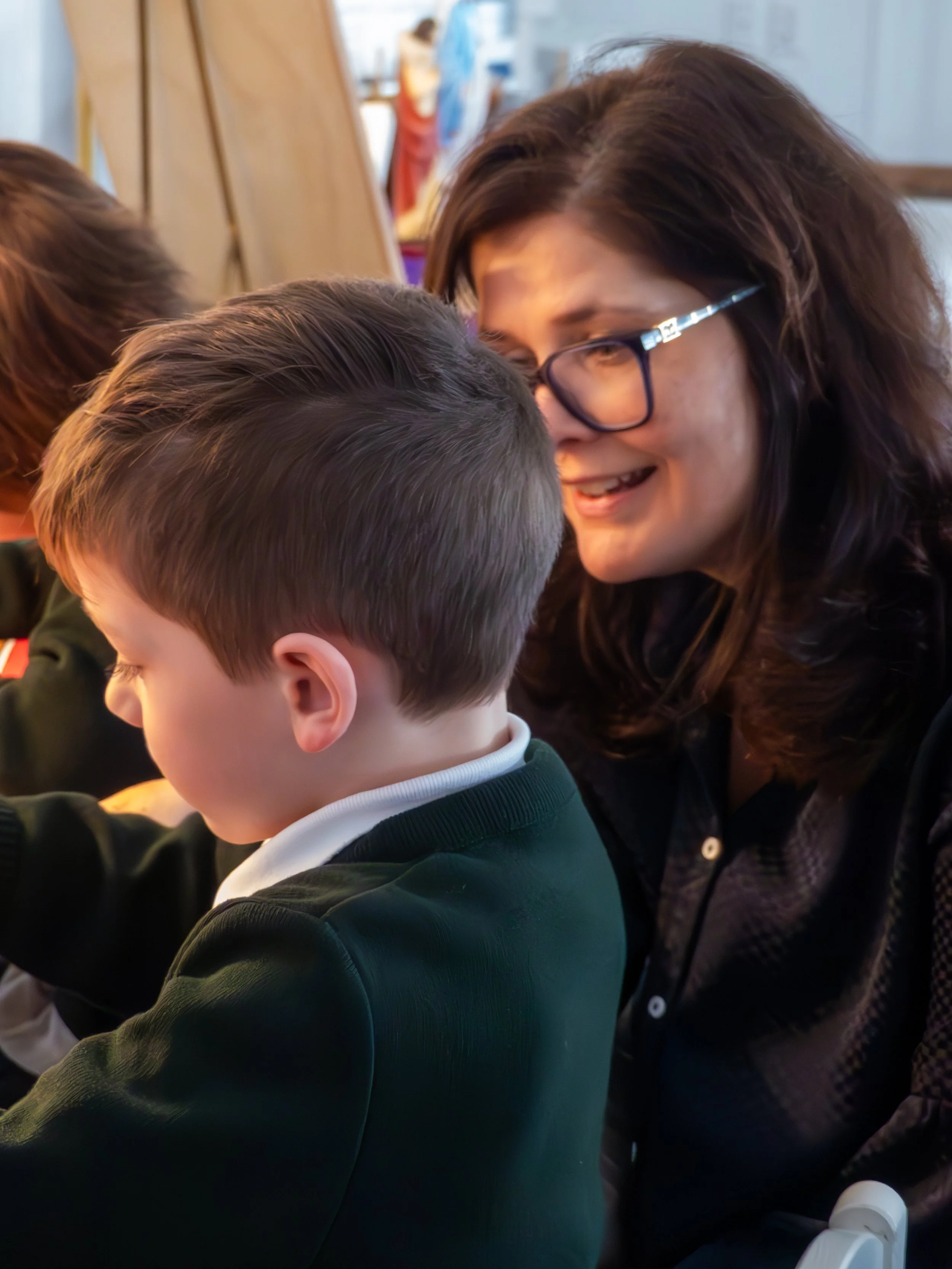 A woman with glasses smiling and leaning close to a young boy with short hair, both engaging with something off-camera in a classroom or similar setting.