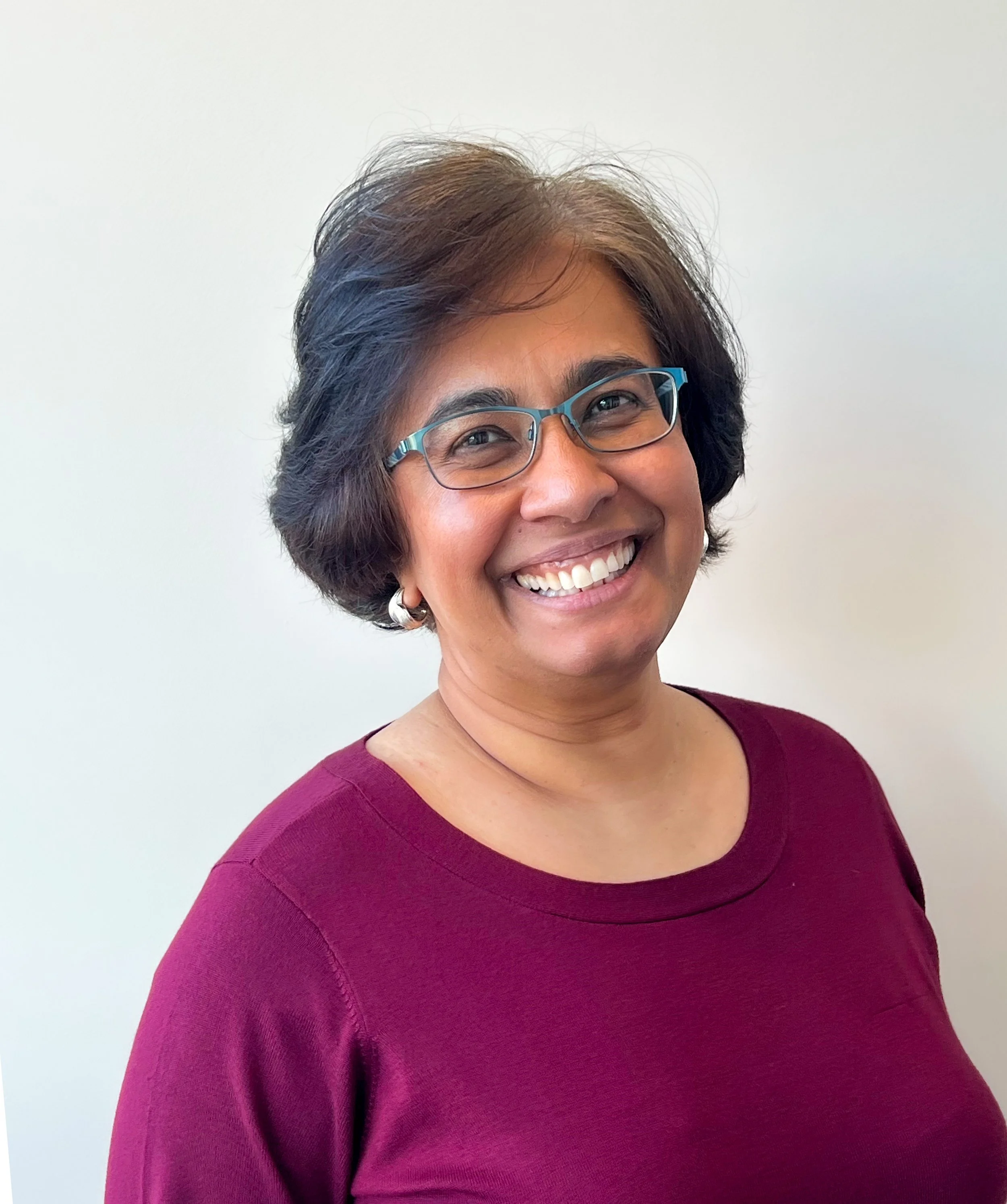 A woman with short dark hair wearing glasses and a maroon top, smiling against a plain white background.