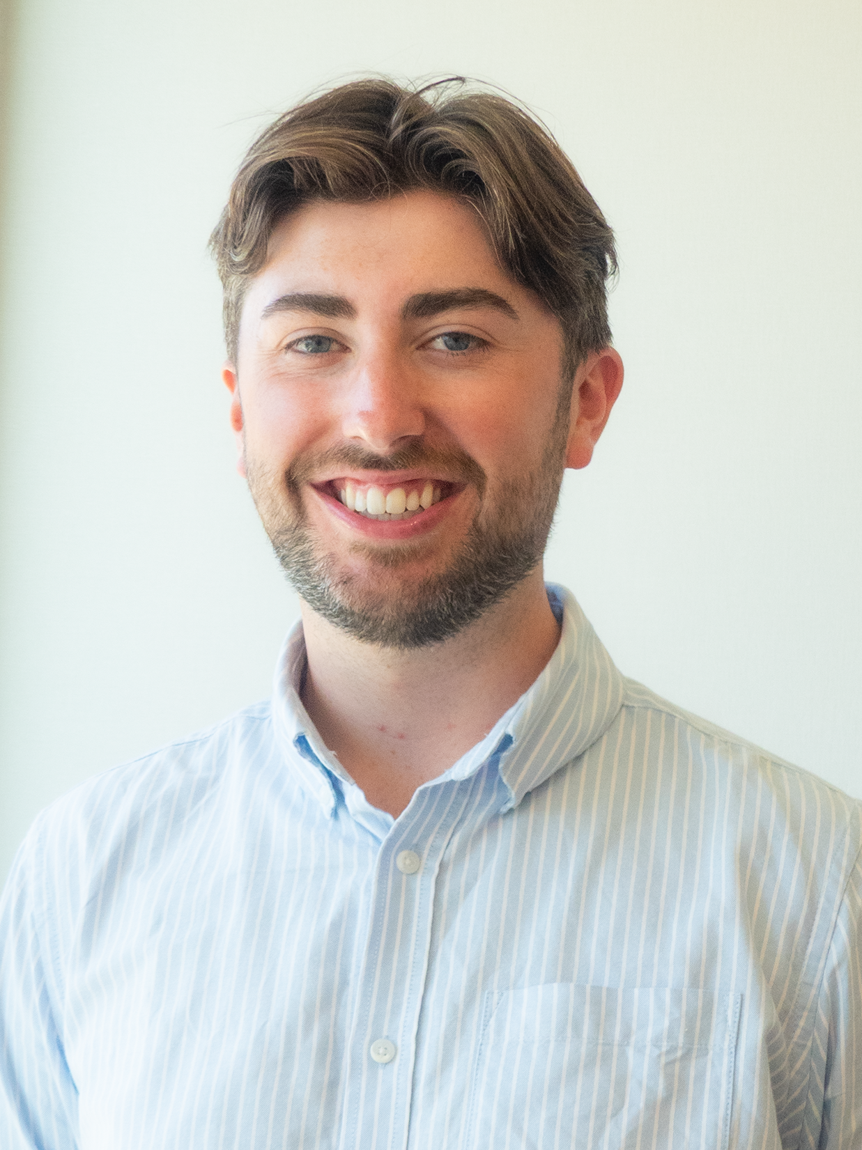 A young man with short dark hair and a beard, smiling, wearing a white collared shirt and a cream-colored sweater, standing against a white wall.