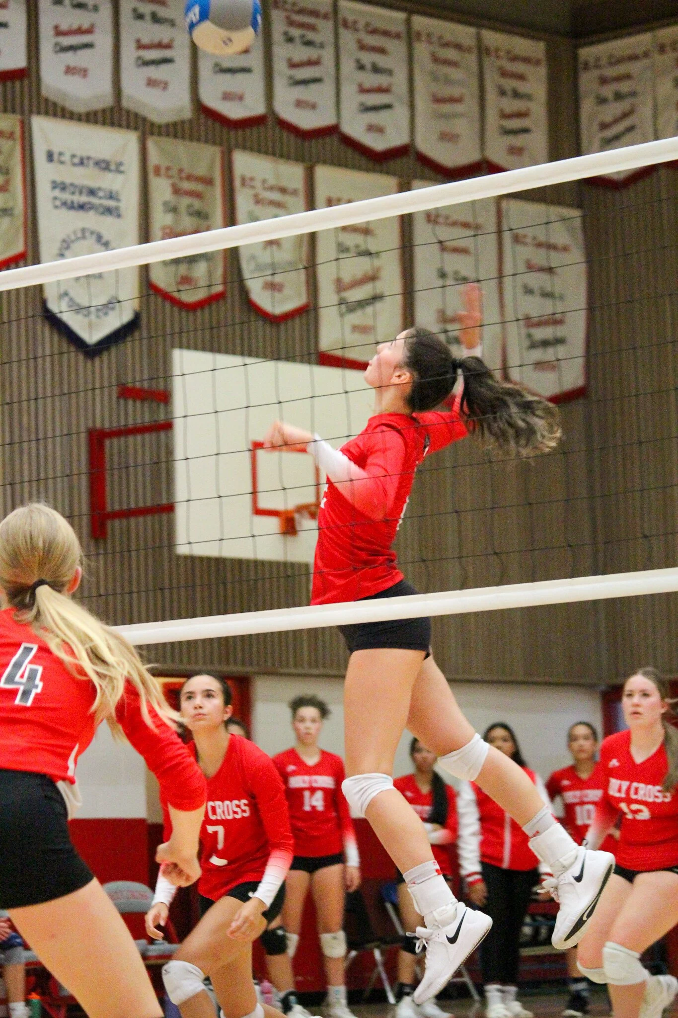 A women's volleyball game in progress inside a gymnasium, with a player in a red uniform jumping to hit the ball over the net while her teammates watch nearby.
