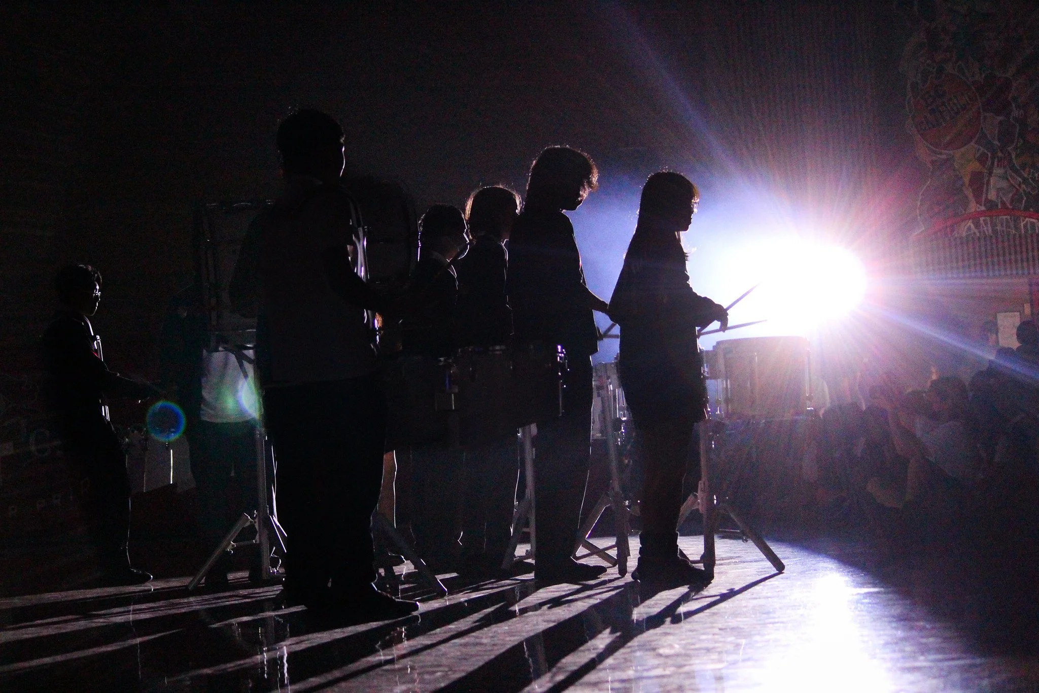 Silhouettes of people playing drums with bright white stage light in the background.