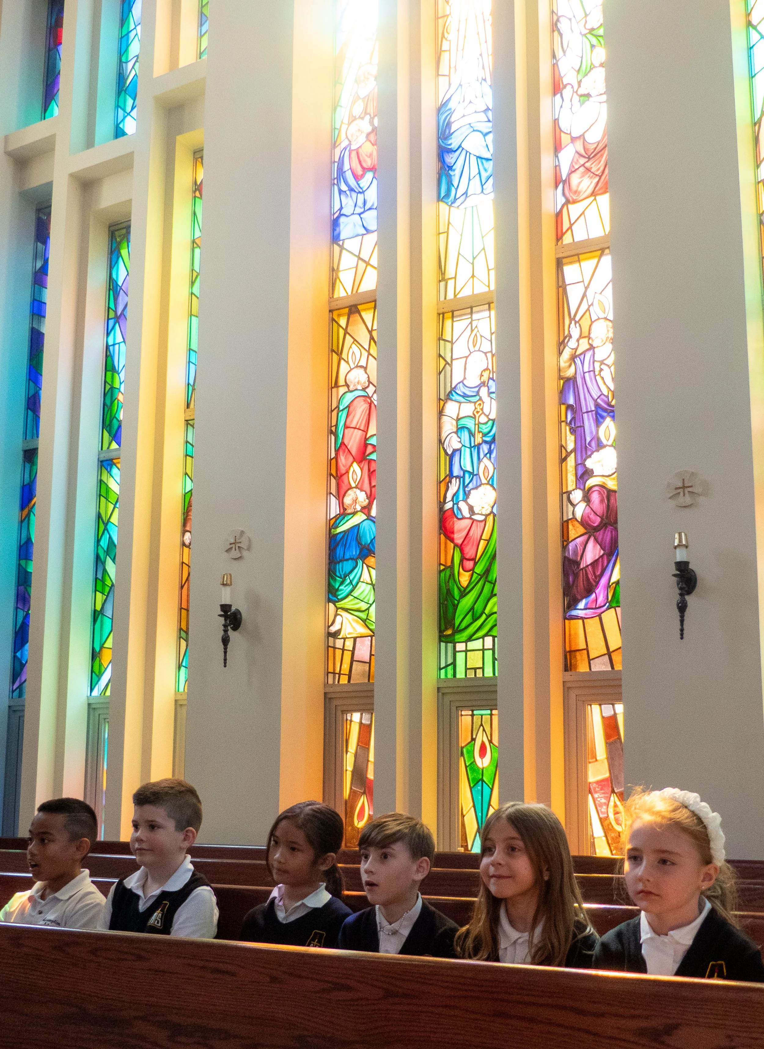 Children sitting in pews inside a church with tall, colorful stained glass windows depicting religious figures and scenes behind them.