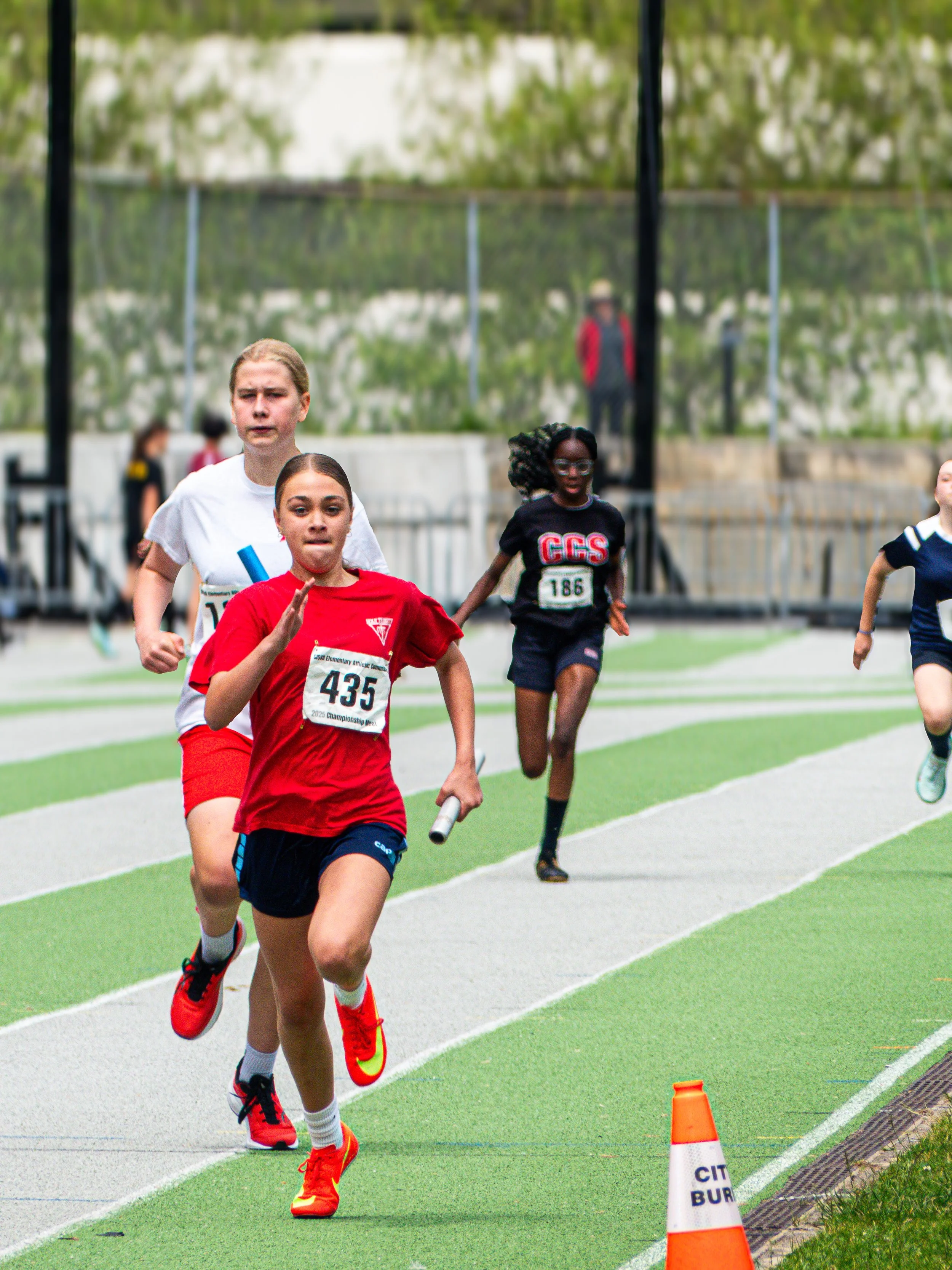 Young girls competing in a running race on a track, one in a red shirt and black shorts, another in a white shirt, and others in dark blue shirts, with a traffic cone on the side.