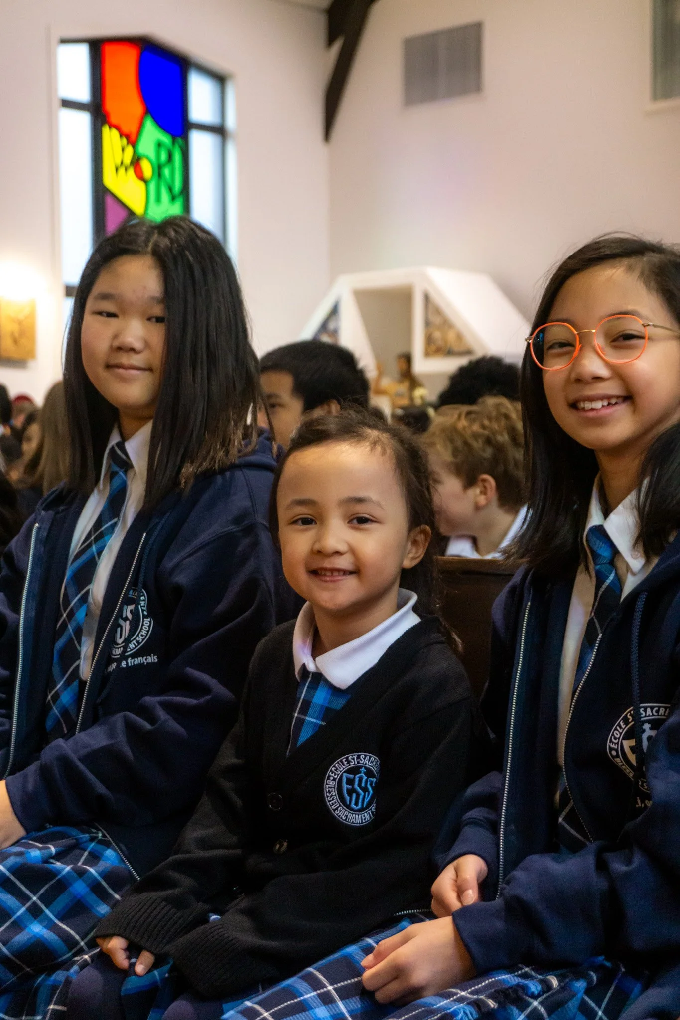 Three schoolgirls in uniforms sitting together inside a church, smiling, with stained glass window behind them.