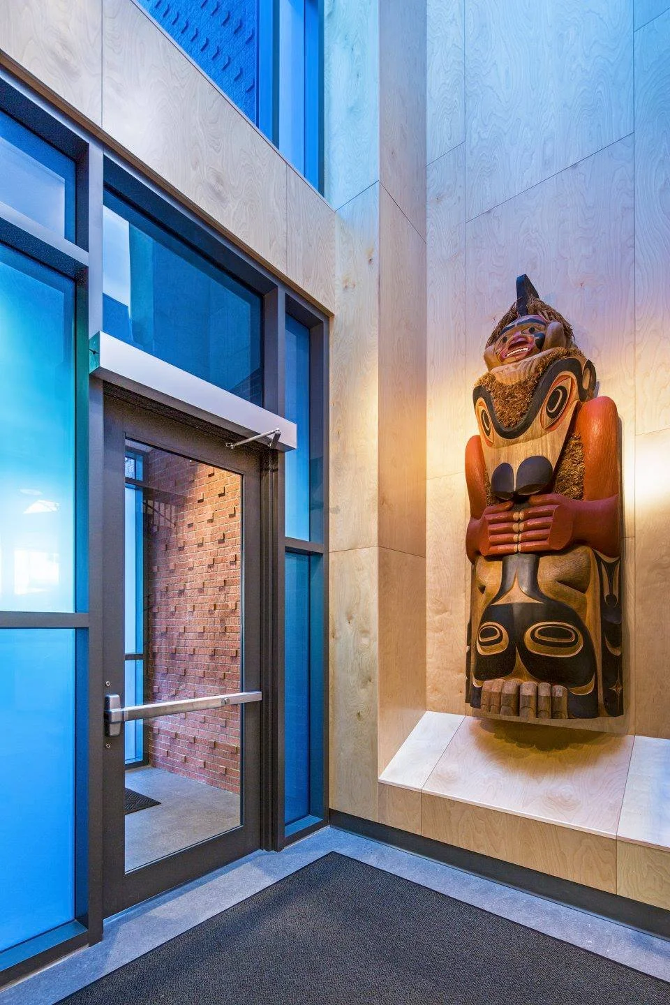 Interior of a building with a decorated wooden Tlingit or Haida style totem pole sculpture mounted on the wall next to a glass door with a brick wall visible outside. The wall inside is covered with light wood panels.