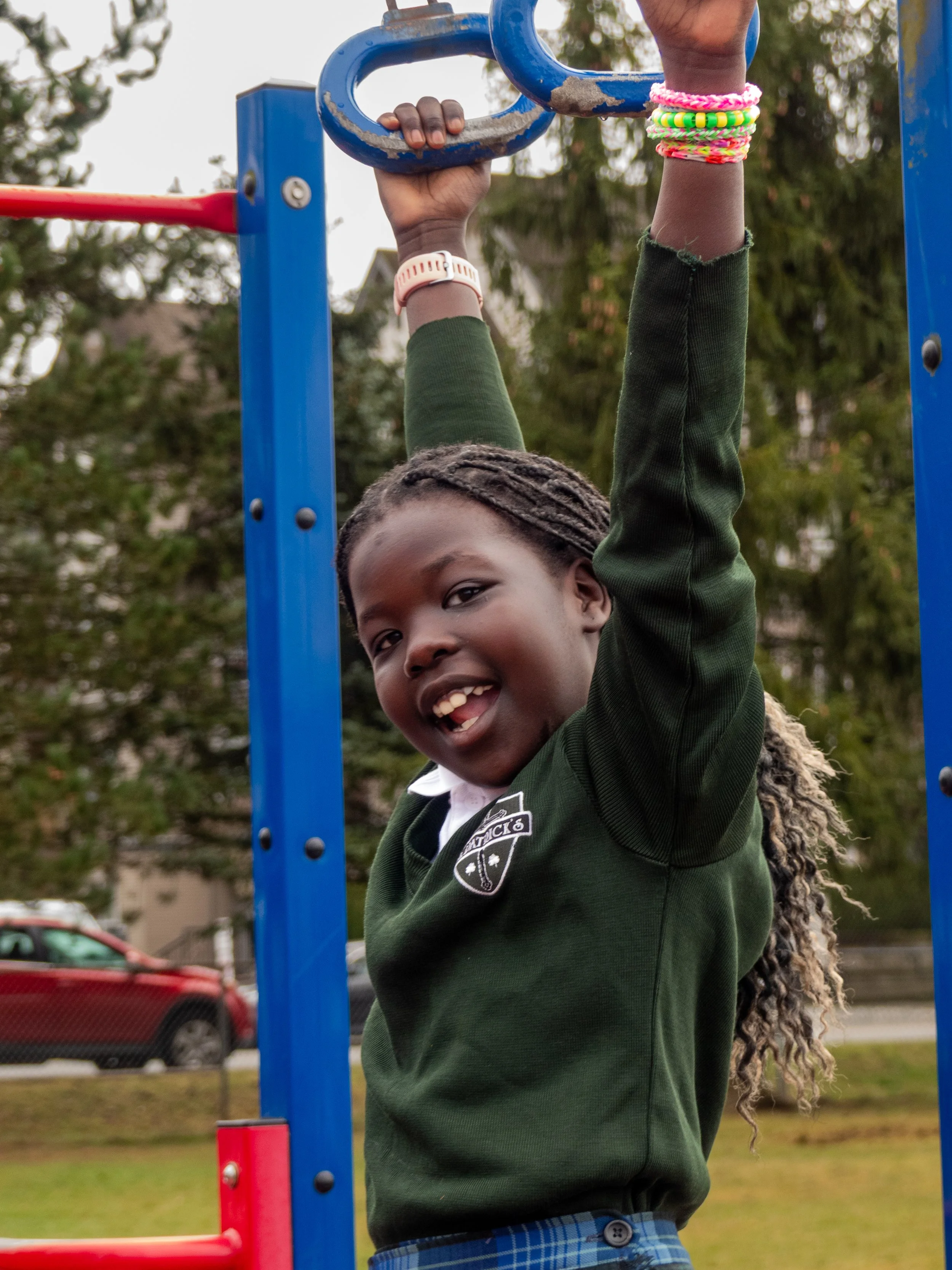 A smiling girl in a school uniform playing on playground equipment, hanging from blue monkey bars with colorful bracelets on her wrist.