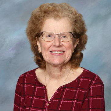 An elderly woman with curly, light brown hair and glasses, smiling, wearing a red plaid top against a soft, multi-colored background.
