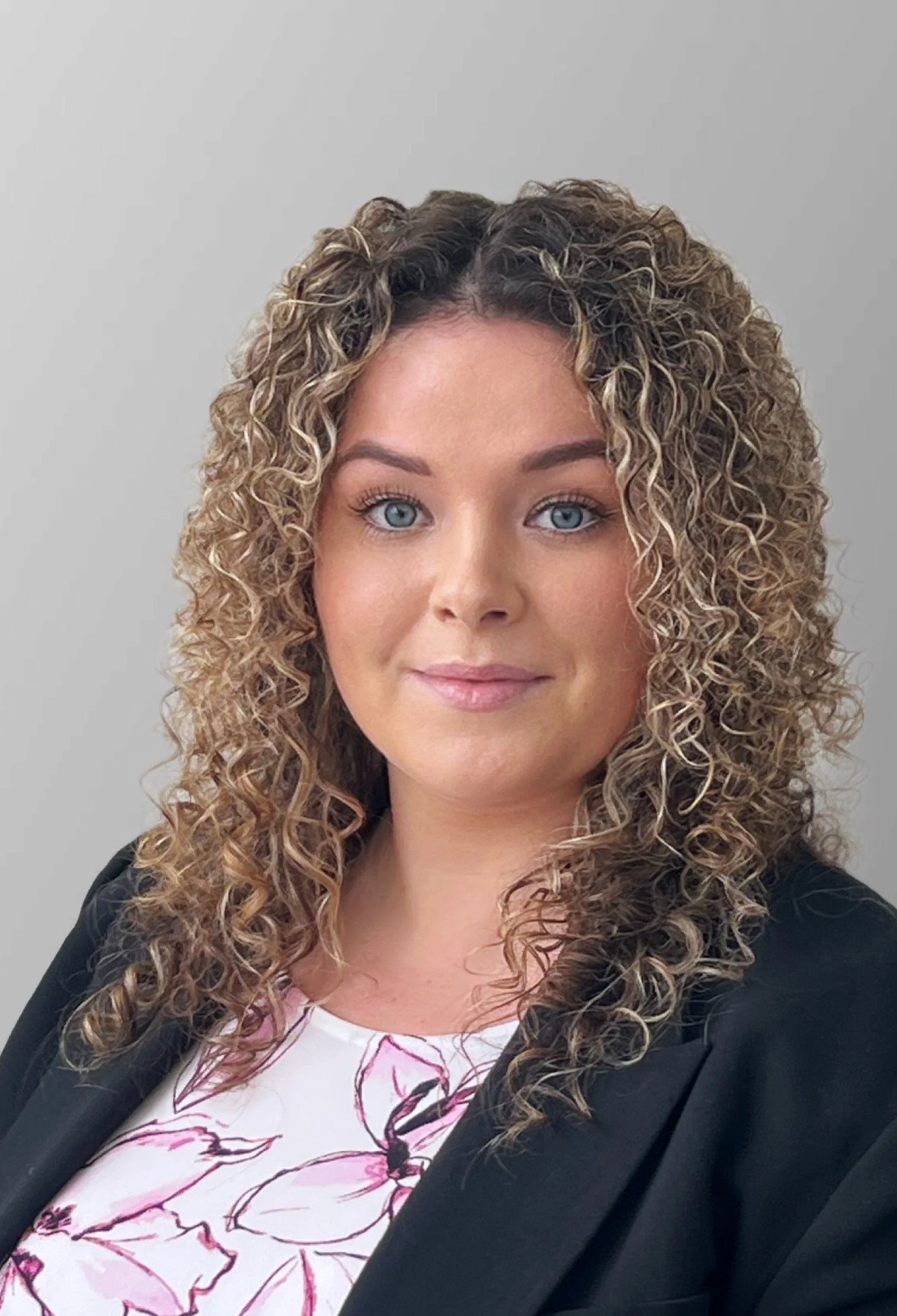 A woman with curly blonde hair wearing a black blazer over a white top with pink floral design, standing against a plain gray background.