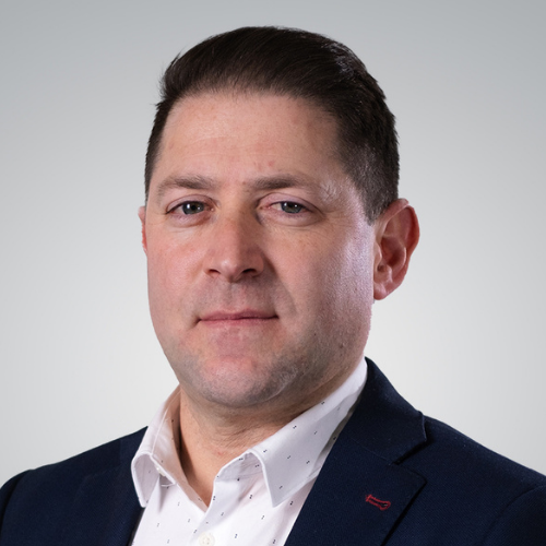 A man with short dark hair wearing a navy blazer and white shirt looking into the camera with a neutral expression.
