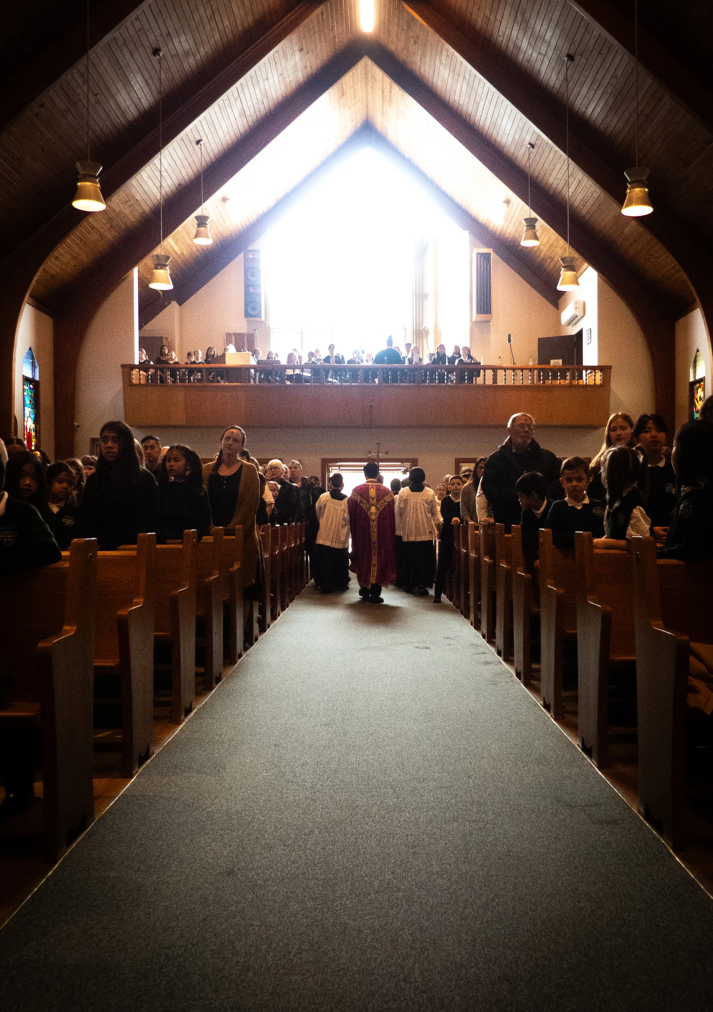 Inside a church during a service with people in pews and altar area, children and adults seated, with a priest in a purple robe near the front, and a bright window at the back illuminating the space.