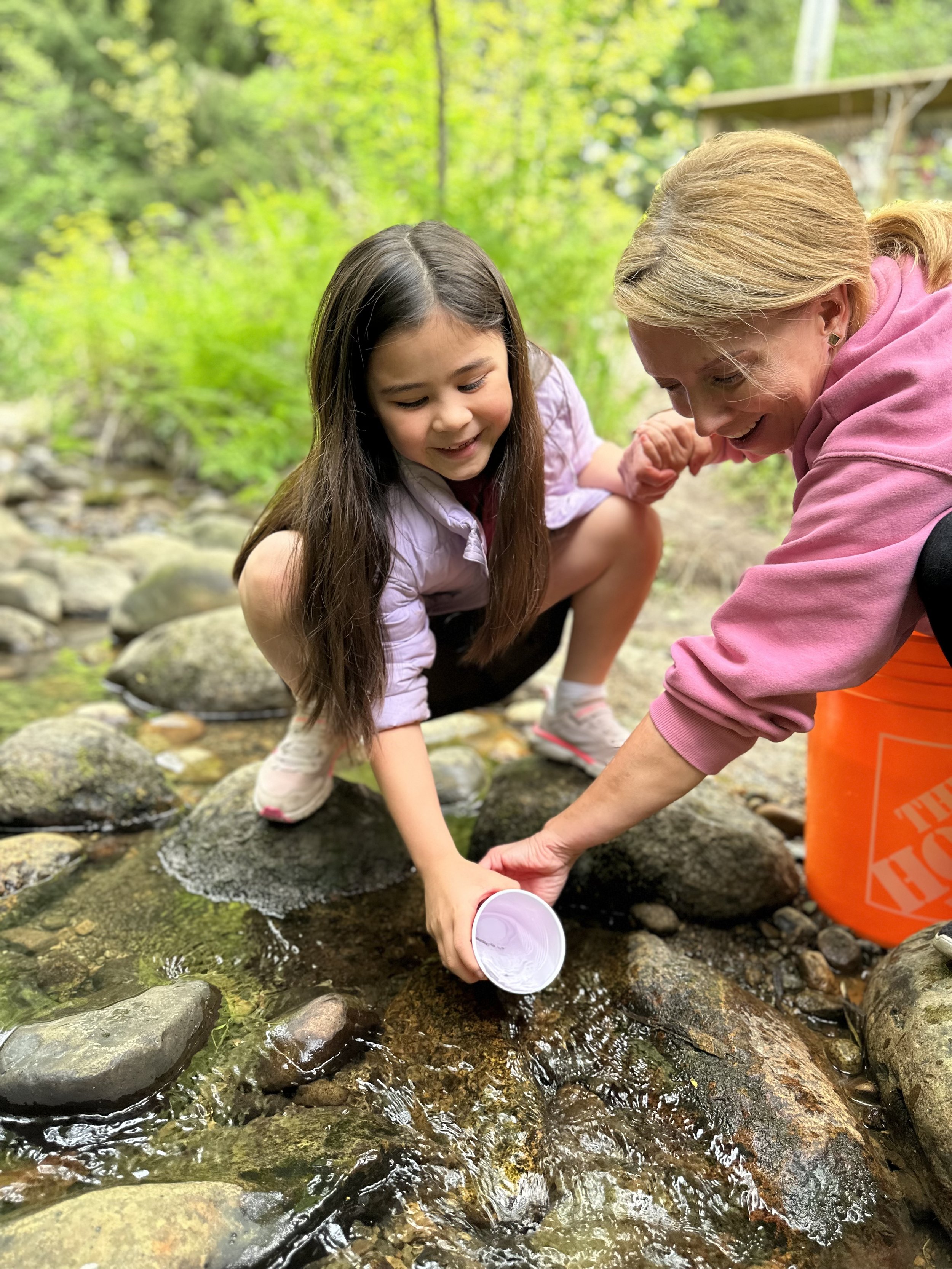 A young girl and an older woman are kneeling on rocks by a creek, smiling and holding a container of water together.