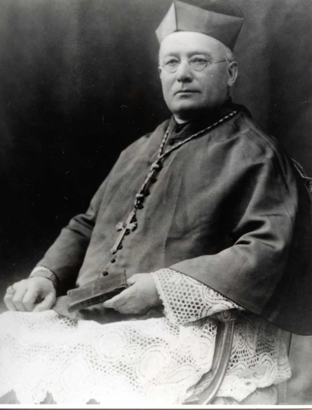 Black and white photograph of a seated man in clerical attire with a cross necklace, holding a small religious book or prayer card, wearing a cap and glasses.