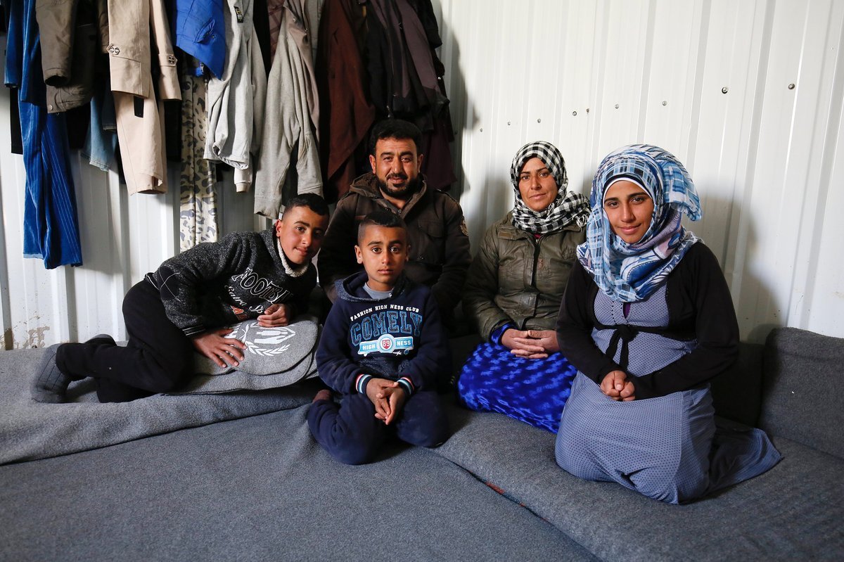 A family of five sitting on a carpeted floor in front of a wall with hanging clothes, including two women wearing headscarves, one man, and two boys.