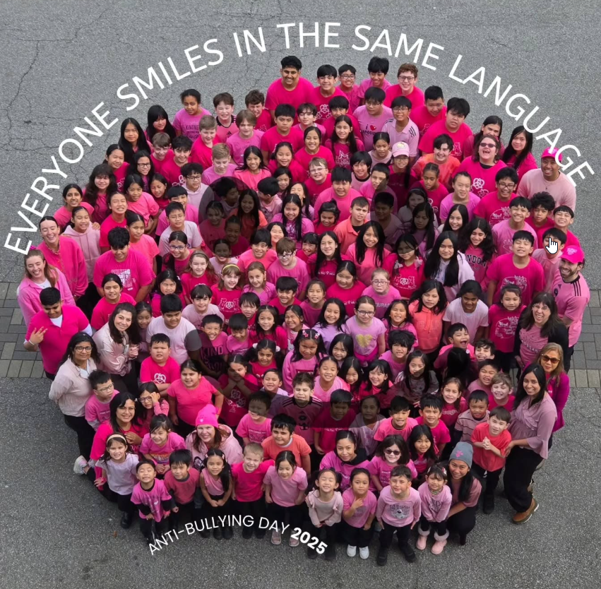 Group of children and adults wearing pink shirts gathered outdoors for Anti-Bullying Day 2025, forming a heart shape, with the caption "Everyone smiles in the same language" written above them.